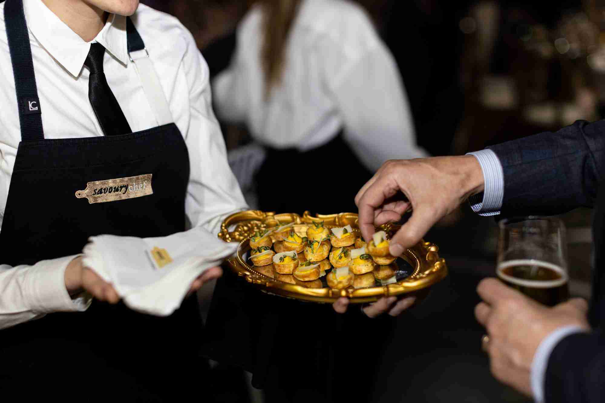 Savoury Chef catering staff serving elegant canapés on a gold platter at a formal event, with a guest reaching for a bite and holding a drink.