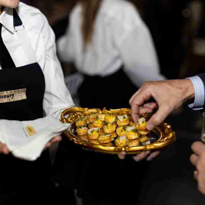 Server in Savoury Chef Catering uniform presenting a tray of gourmet appetizers at an event, with a guest reaching for a bite-sized treat.